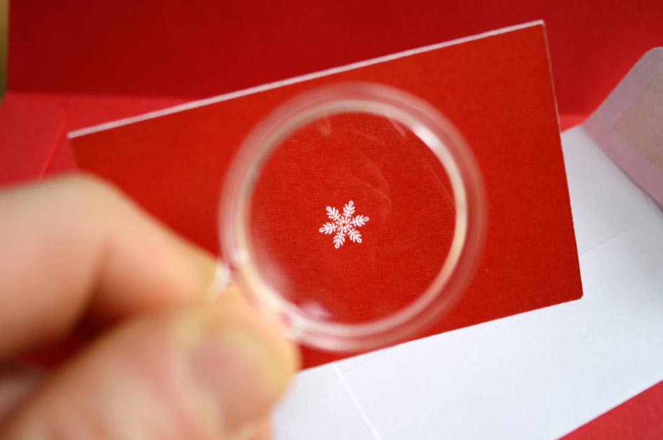 A magnifying glass shows a closeup of a snowflake printed on the front of a tiny red holiday card.
